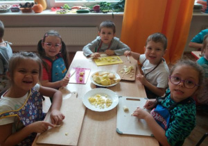 dzieci w fartuszkach siedzą przy stoliku i na deskach kroją jabłka a na środku stolika stoją dwa talerze z kawałkami jabłek i drugi z pokrojonymi na małe kawałki